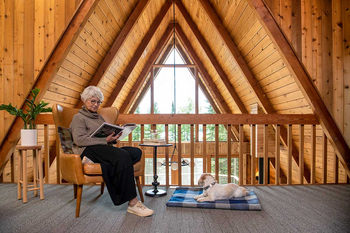 A woman reading a book in a leather chair next to a dog laying on a blanket on the floor in front of Marvin Ultimate triangular windows in an A-frame cabin in Northern Minnesota.